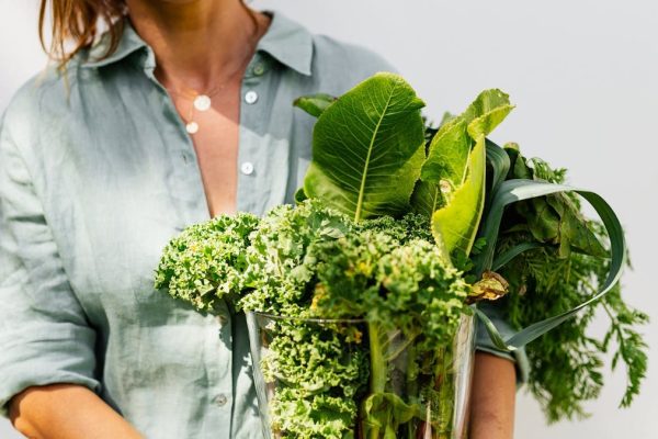 A woman in a light shirt carrying a basket of fresh greens, symbolizing healthy eating and lifestyle.